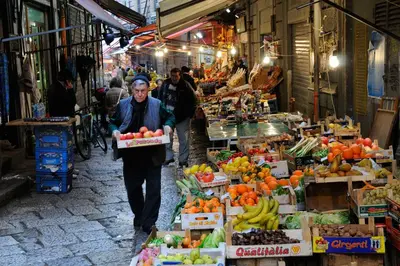 a woman walking through a market with fruits and vegetables at Caleidoscopica Bahlara Ballarò in Palermo