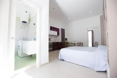 a white bedroom with a white bed and a kitchen at Casa in centro in Castellammare del Golfo