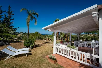 a white house with a porch and a chair and a palm tree at NUOVA Villa Intera Guidaloca in Scopello