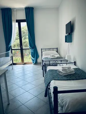 a room with three beds and a window with blue curtains at Hotel Achibea in San Vito lo Capo